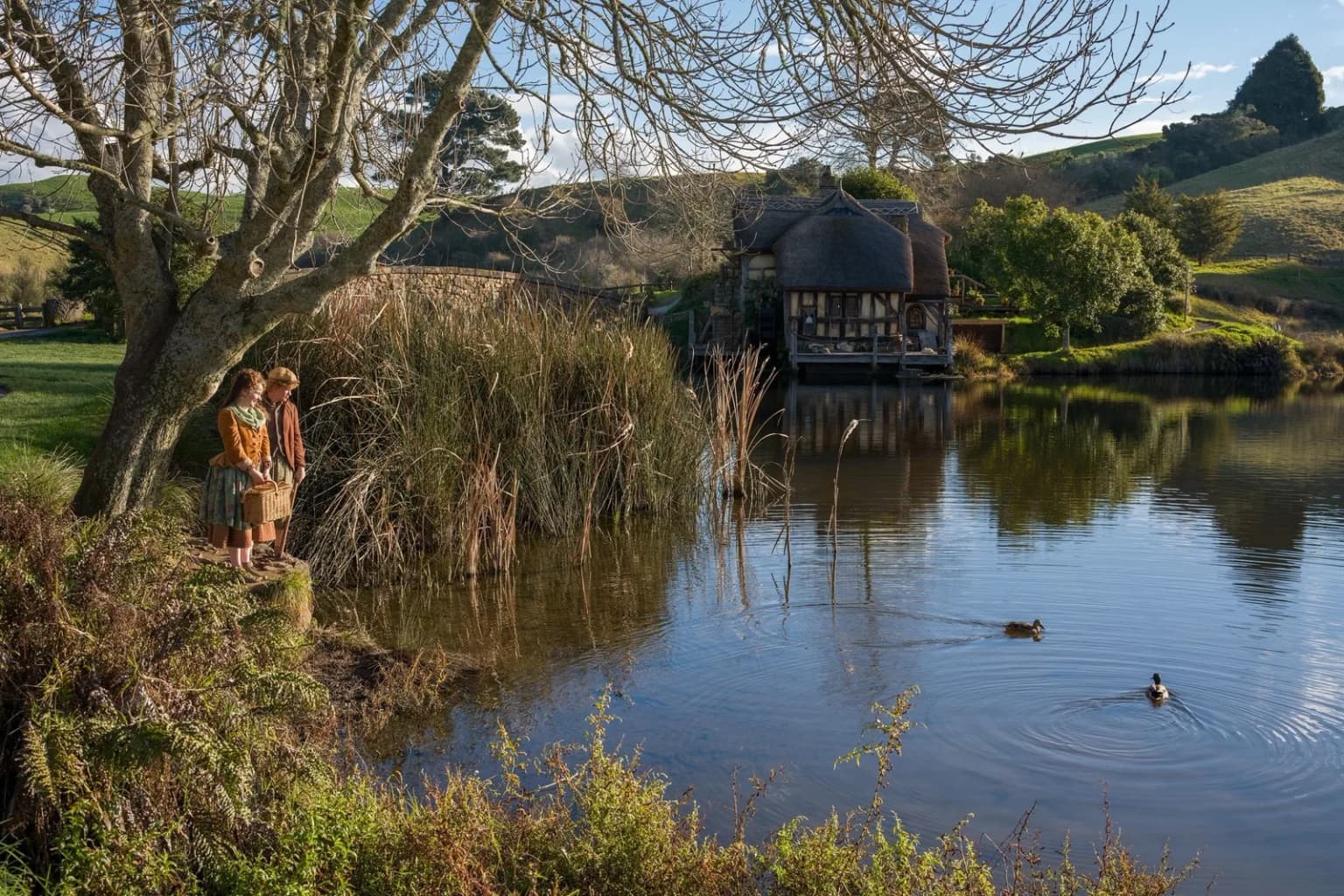 Deux jeunes Hobbits observent des canards au bord de l'étang, avec le Moulin de Hobbiton en arrière-plan