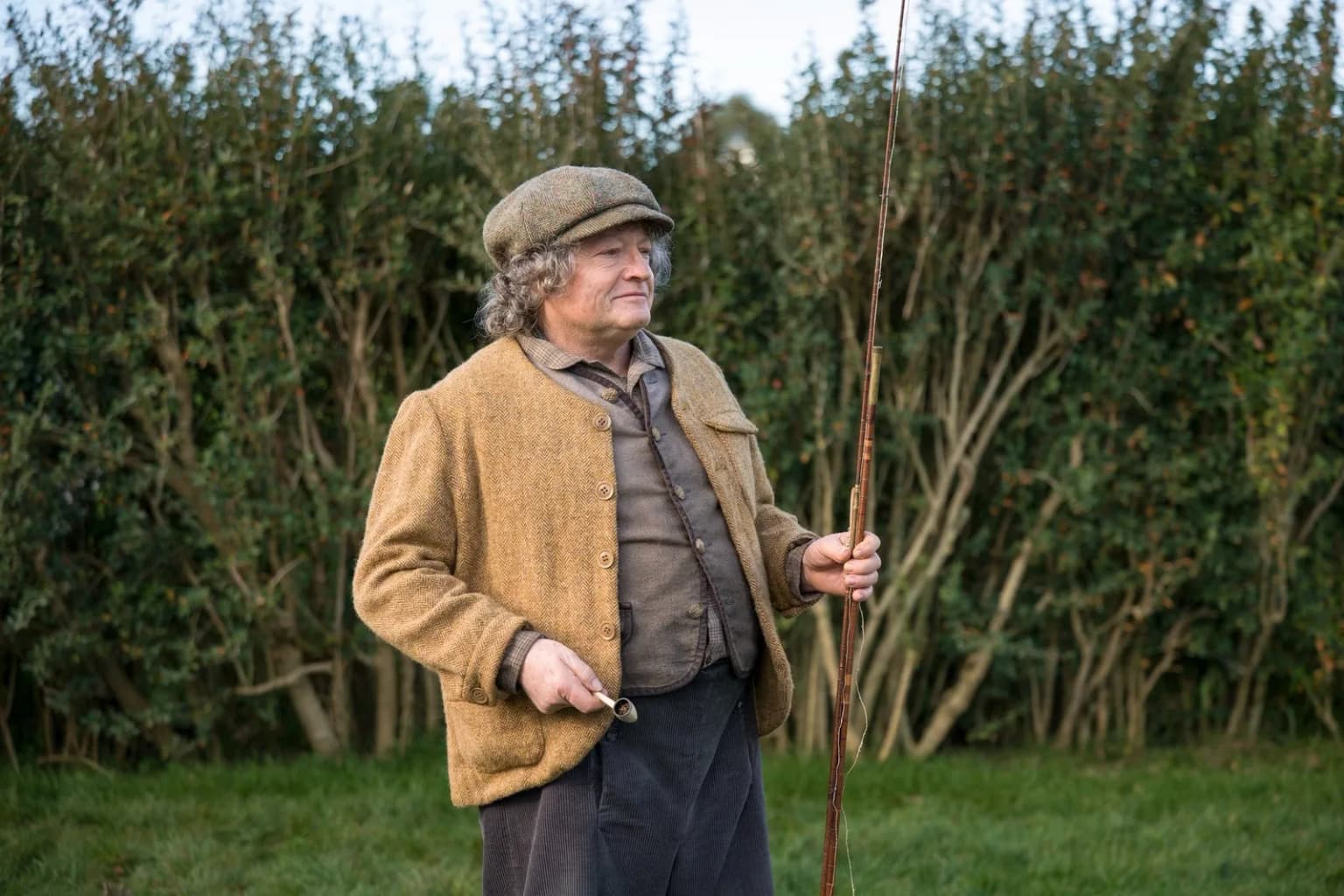 Un vieil Hobbit avec casquette et canne à pêche pose devant une haie